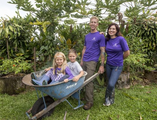 Founders of Hoʻōla Farms in Hilo, Hawaii, with their two daughters in a wheelbarrow on the farm. Husband holds the wheelbarrow while his wife stands beside him, representing family and community farming.